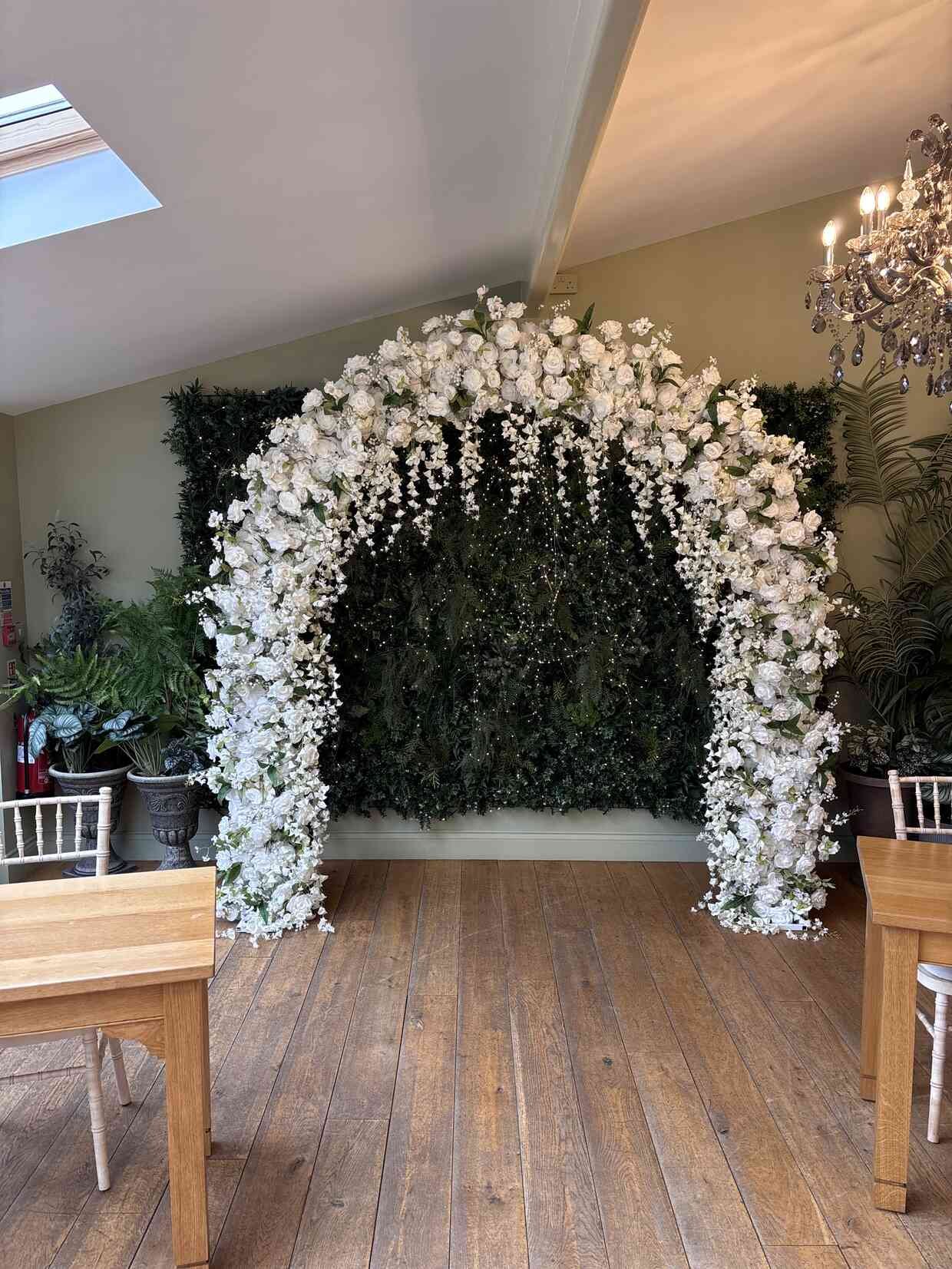 White floral arch with cascading roses set against a green flower wall with fairy lights — wedding venue